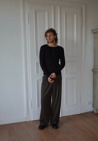 Camicia nera a maniche lunghe con bottoni, abbinata a pantaloni larghi marroni a righe sottili e scarpe nere. Pavimenti in legno e porte bianche a pannelli sullo sfondo.