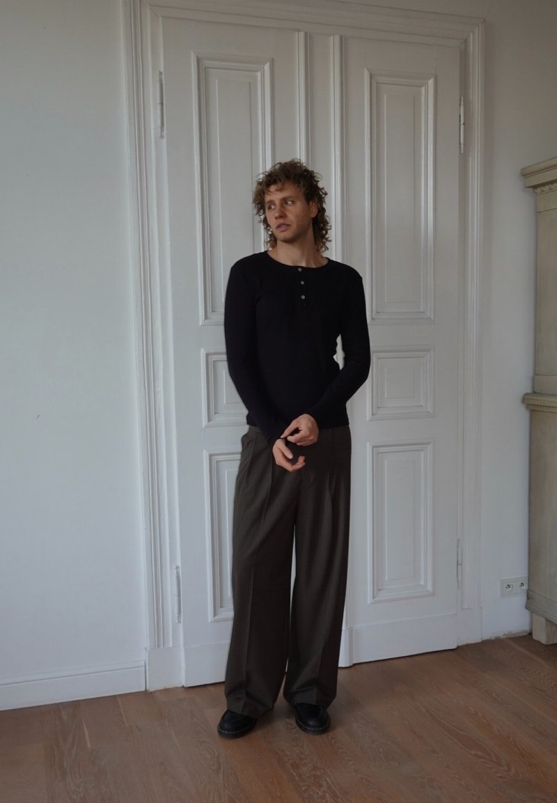 Camicia nera a maniche lunghe con bottoni, abbinata a pantaloni marroni larghi a righe sottili e scarpe nere. Pavimenti in legno e porte bianche a pannelli sullo sfondo.