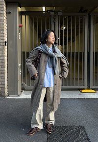 Jeune personne debout, les mains dans les poches, portant un manteau à carreaux, une chemise bleue, un pantalon beige, un pull gris noué autour des épaules, des lunettes de soleil sur la tête.