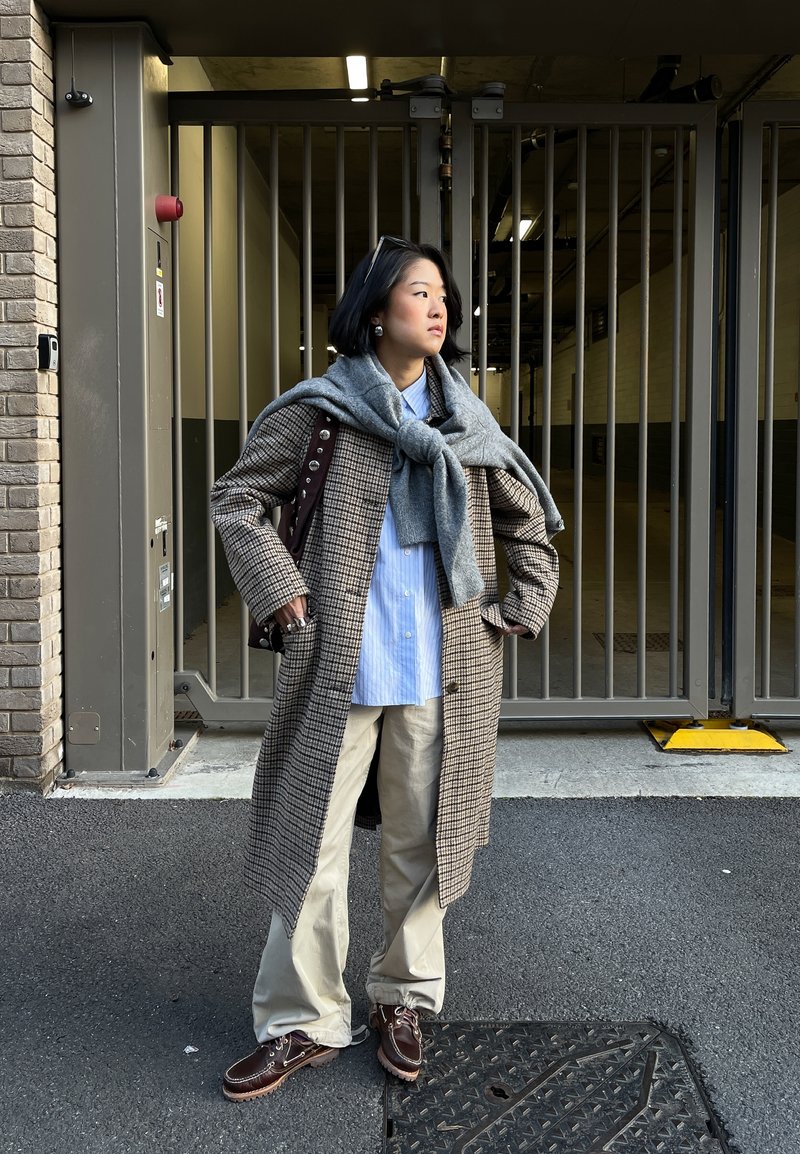 Jeune personne debout, mains dans les poches, portant un manteau à carreaux, une chemise bleue, un pantalon beige, un pull gris noué autour des épaules, des lunettes de soleil sur la tête.