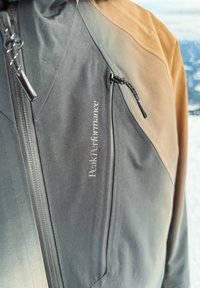 Δίχρωμο μαύρο και καφέ μπουφάν Peak Performance με εμφανείς φερμουάρ και τσέπη στο στήθος σε εξωτερικό χιονισμένο τοπίο.