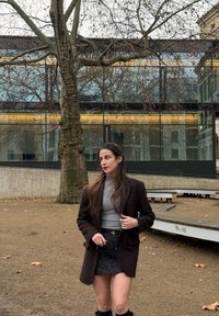 Νεαρή γυναίκα με σκούρο σακάκι και γκρι μπλούζα ζιβάγκο στέκεται σε εξωτερικό χώρο κοντά σε δέντρο χω ρίς φύλλα και σύγχρονο γυάλινο κτήριο σε μια συννεφιασμένη μέρα.