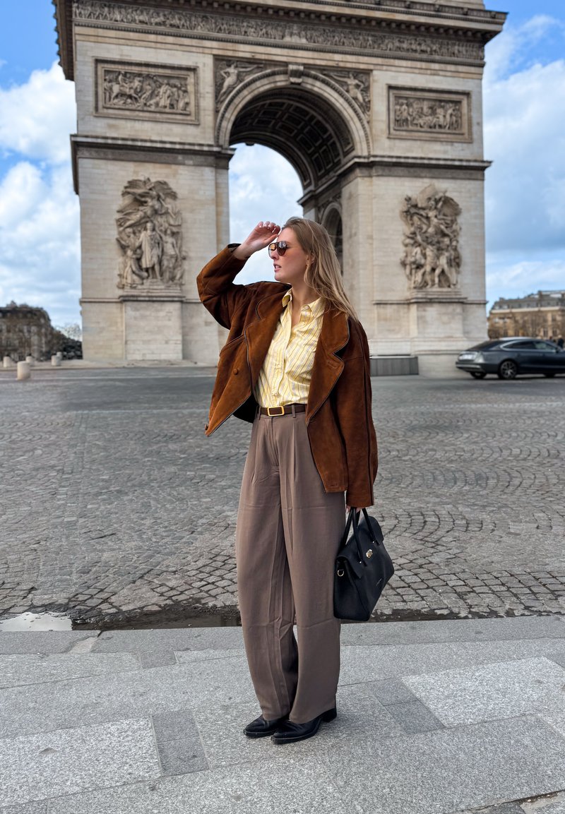 Femme portant une veste marron, une chemise jaune et un pantalon large, tenant un sac à main noir, debout près de l'Arc de Triomphe à Paris.