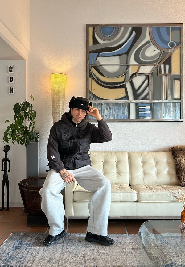 Musta työliivi harmaan hupunpaidon päällä, harmaat verkkarit ja kiiltävät mustat kengät. Istuu kermanvärisellä sohvalla, jonka ympärillä on taidetta ja sisustusesineitä.