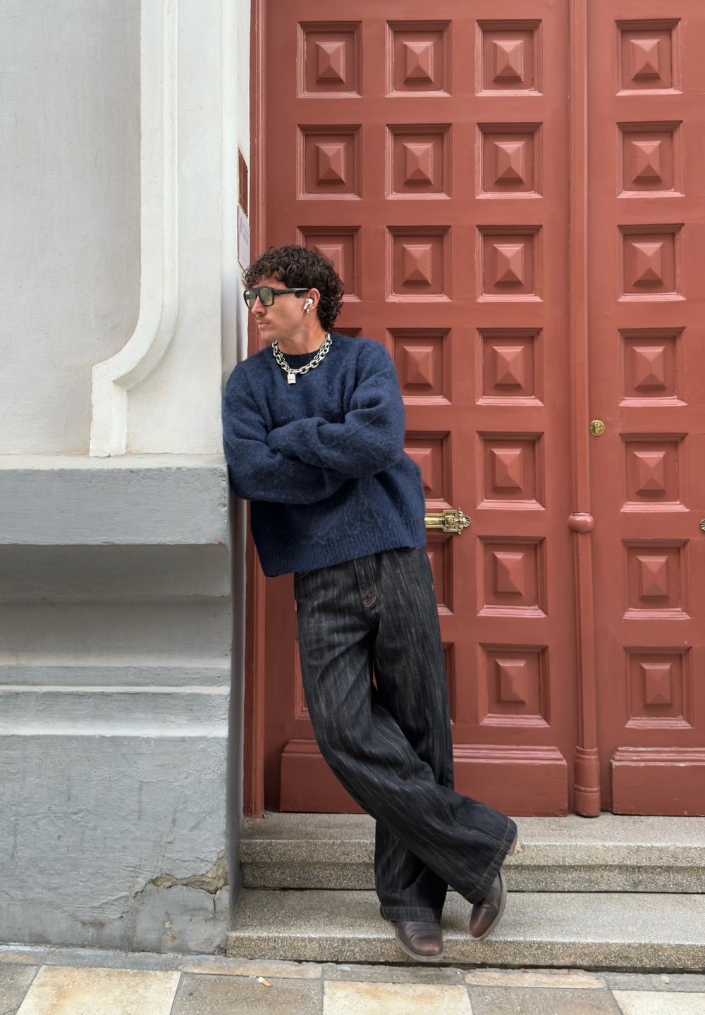 Maglione blu scuro in peluche, jeans a gamba larga scuri, collana a catena spessa, occhiali da sole e AirPods, appoggiato a un muro testurizzato accanto a una porta di legno.