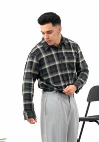 Chemise à carreaux noirs et verts avec un col boutonné, des manches longues et une poche avant, associée à un pantalon gris clair et une ceinture cloutée.