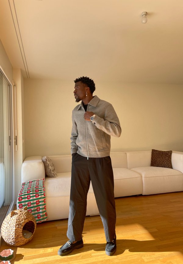 Man wearing grey jacket and black pants stands inside living room with white sofa and wicker pet basket nearby.