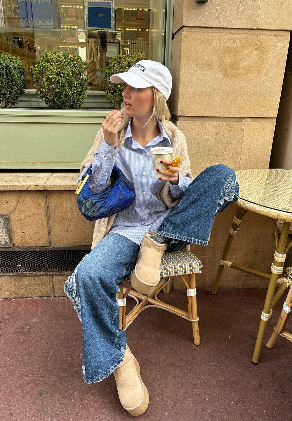 Μπλε καρό πουκάμισο, μπεζ ζακέτα, μπλε τσάντα, σκισμένα μπλε τζιν, κρέμα UGG μπότες. Κρατάει ένα ποτό και ένα σνακ ενώ κάθεται.