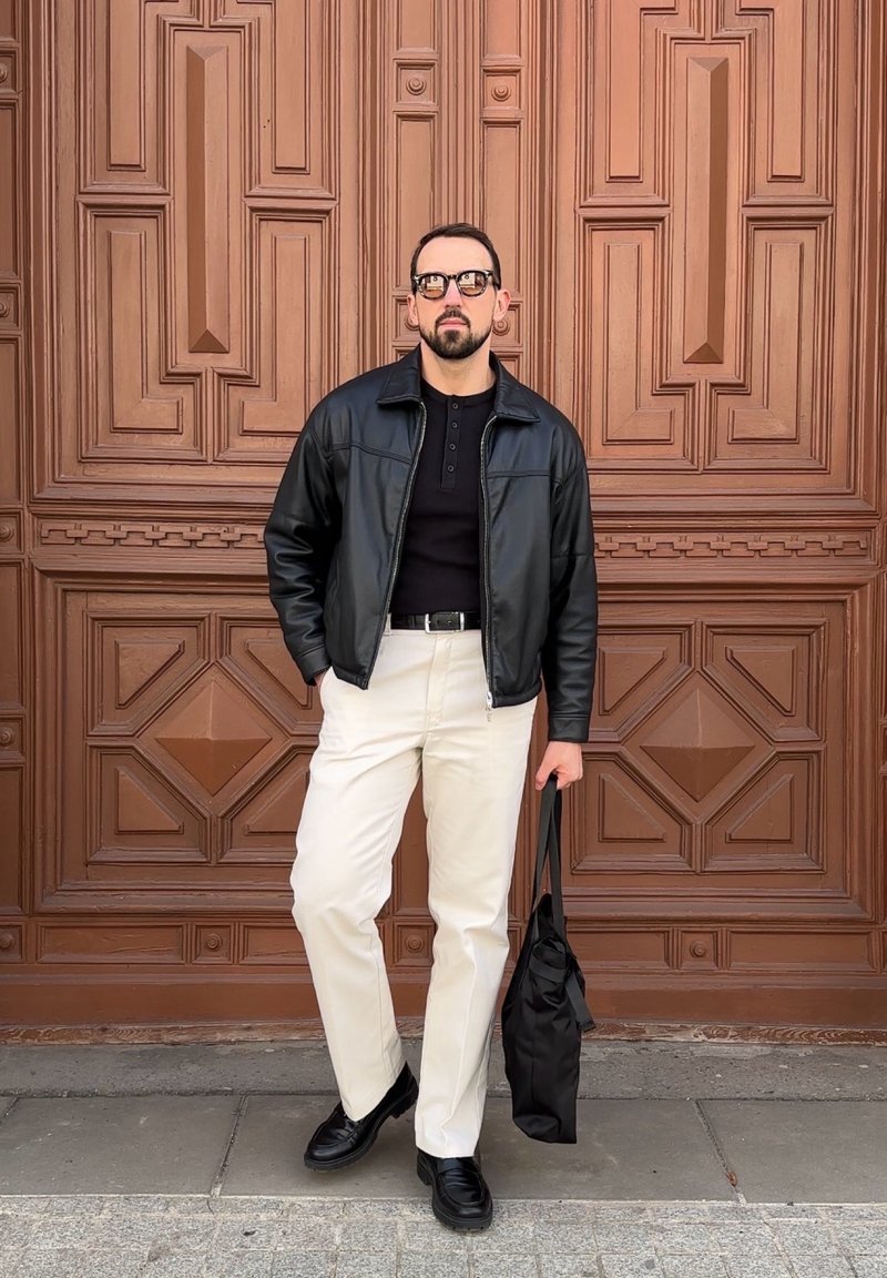 Homme portant une veste en cuir noire, une chemise noire, un pantalon blanc, des chaussures noires et des lunettes, se tient avec un sac cabas noir devant une porte en bois sculpté.