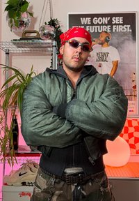 Zöld bomberdzseki párnázott textúrával és fekete részletekkel, fekete felső fölött viselve. A megjelenést terepmintás nadrág és piros bandana egészíti ki.