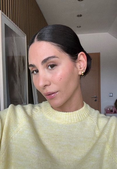 Suéter de punto amarillo claro con un cuello acanalado, textura suave. Cabello castaño recogido en un moño bajo, piel radiante, aretes de oro, fondo neutro.