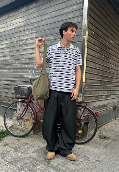 Camisa polo de manga corta a rayas, pantalones de pierna ancha negros, bolso de hombro verde oliva, zuecos tostados, con una bicicleta de fondo.