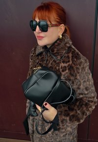 Sac en cuir noir avec une fermeture éclair dorée tenue dans une main. La personne porte un manteau marron à imprimé léopard avec un col haut et de grandes lunettes de soleil noires.