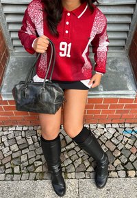 Rdeče in bele vzorčaste polo s dolgimi rokavi z številko 91, v kombinaciji z črno usnjeno torbico in črnimi škornji do kolen.