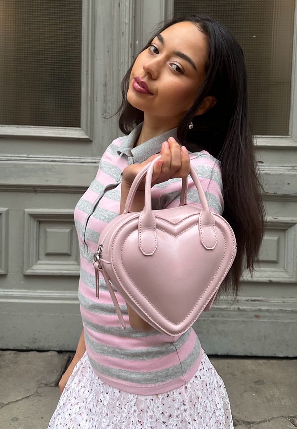 Junge Frau hält eine pinke, herzförmige Handtasche vor die Kamera, trägt ein gestreiftes Hemd und einen Spitzenrock, steht vor einer Holztür.