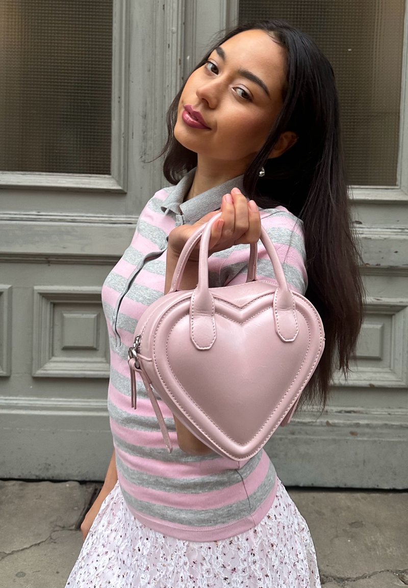Jeune femme tenant un sac à main rose en forme de cœur vers la caméra, portant une chemise rayée et une jupe en dentelle, debout devant une porte en bois.