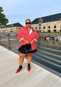 Red and white striped oversized shirt, black shorts, red pointed heels, and black sunglasses. Accessories include a pearl necklace and a black bag.