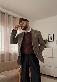 Άνδρας που φοράει καφέ σκουφί, γυαλιά, καρό σακάκι, καφέ πουλόβερ και μαύρο παντελόνι σε ένα φωτεινό σαλόνι με μπεζ κουρτίνα και λευκή ντουλάπα.