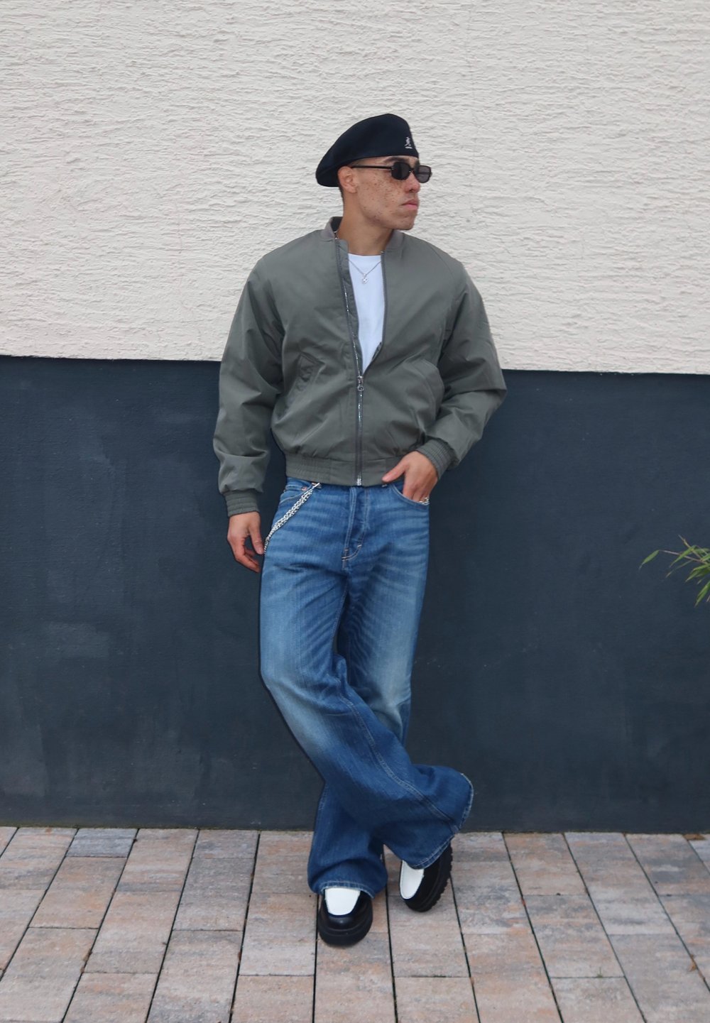 Casaco bomber verde-oliva, t-shirt branca, jeans azuis amplos, sapatos pretos com detalhes brancos, boina preta e óculos de sol. Fundo de parede texturizada.