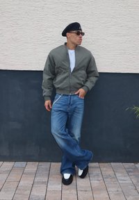 Casaco bomber verde-oliva, t-shirt branca, jeans azuis amplos, sapatos pretos com detalhes brancos, boina preta e óculos de sol. Fundo de parede texturizada.