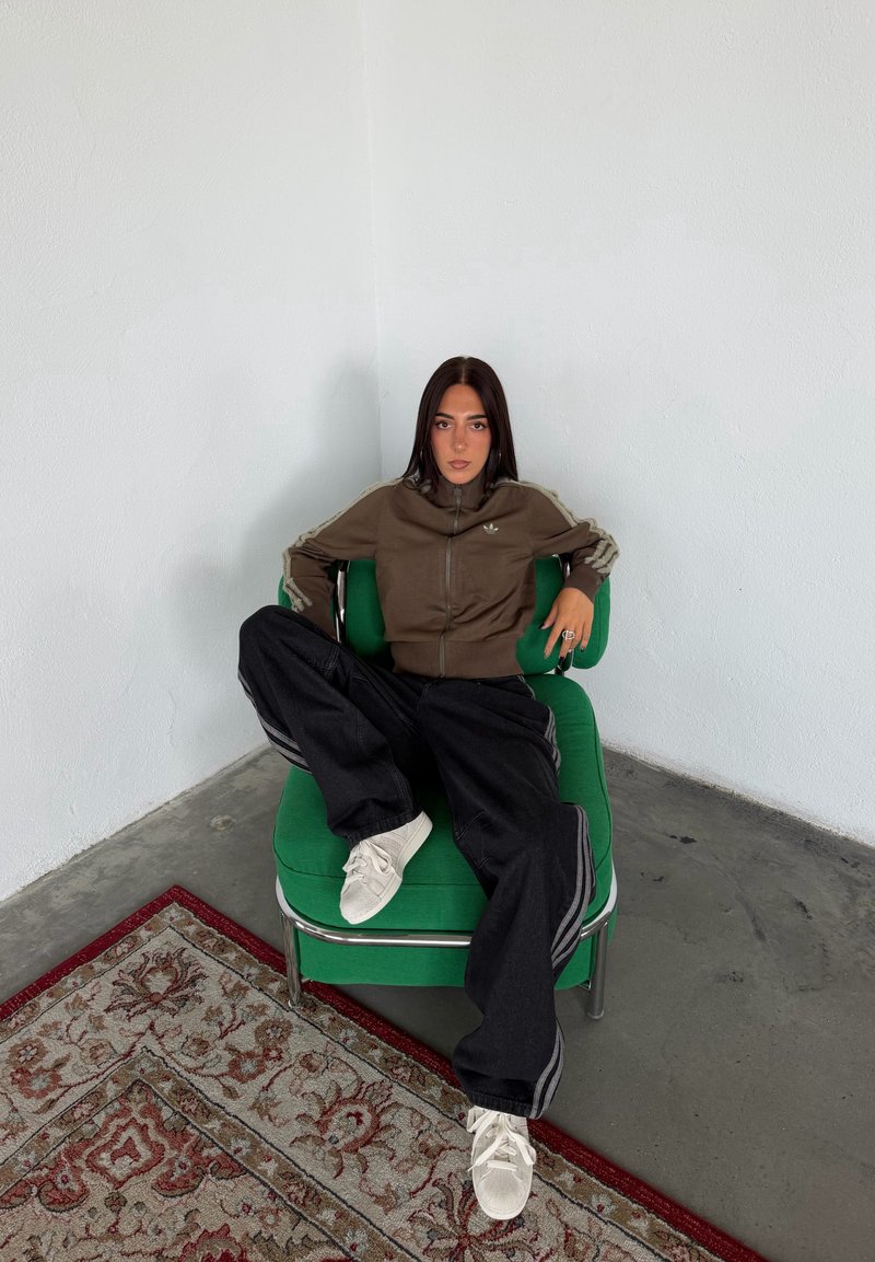 Femme aux longs cheveux foncés assise décontractée sur une chaise verte dans un coin, portant une veste marron, un pantalon noir et des baskets blanches.