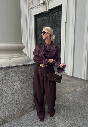 Braune Satinbluse mit Rüschen, hochgeschnittene braune weite Hose und eine braune Handtasche. Accessoires umfassen eine Blumenbrosche und Sonnenbrille.