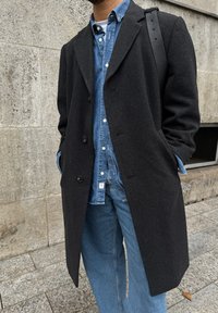 Μαύρο μάλλινο παλτό με εγκοπές, μονό κουμπί, συνδυασμένο με τζιν πουκάμισο και φαρδιά μπλε τζιν.