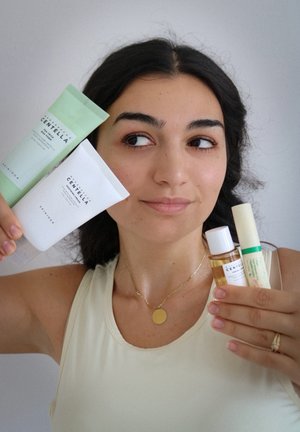 Tre prodotti per la cura della pelle: un tubo verde, un tubo bianco, una piccola bottiglia marrone e un bastoncino bianco con un accento verde, tenuti da una persona.