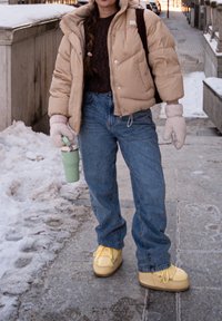 Personne en doudoune beige, jean et chaussures d'hiver jaunes tenant un gobelet de voyage vert, debout sur un trottoir enneigé en milieu urbain.