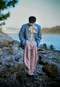 Hele helesinine bleiser, valge mustriga särk, suur valge kikilips, roosa pükste ja heledad jalanõud; seisab kivisel rannikul vee lähedal.