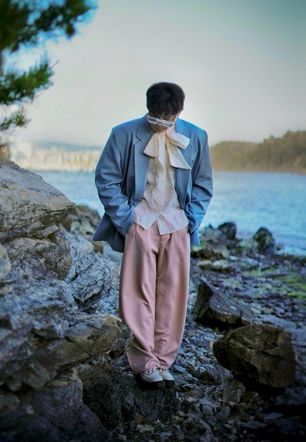 Blazer azul claro, camisa blanca con estampado, gran pajarita blanca, pantalones rosas y zapatos claros; de pie en una costa rocosa cerca del agua.