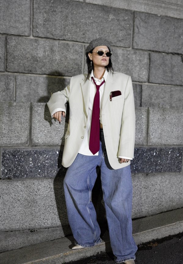 Persona que lleva un blazer crema extragrande, camisa blanca, corbata roja, vaqueros azules anchos, boina y gafas de sol apoyada en una pared de piedra al aire libre.