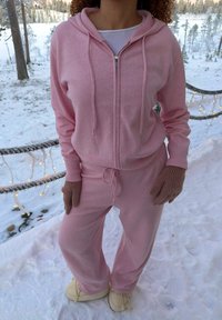 Persoon die een roze hoodie en joggingbroek draagt, bruine handschoenen en crème schoenen, staand in een besneeuwde buitenomgeving met bomen en lichtslingers.