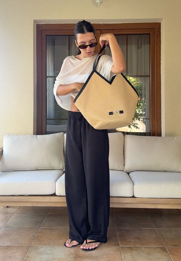 Frau mit Sonnenbrille und schwarzer Hose schaut in eine große, geflochtene Tragetasche, während sie auf einem gefliesten Boden in der Nähe eines weißen Sofas steht.