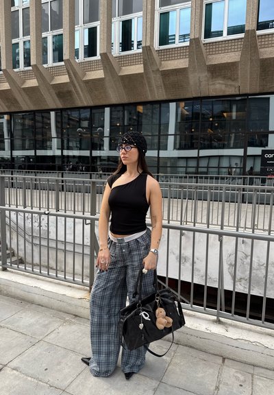 Top sin mangas negro, pantalones anchos de cuadros grises, bolso negro con un charm de oso de peluche y gafas redondas, con fondo de un edificio de cristal moderno.
