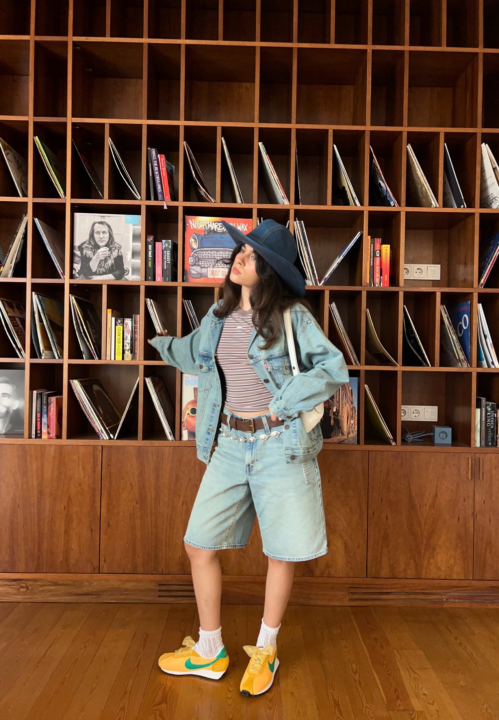 Jeansjakke, stripete skjorte, lyse denimshorts og gule sneakers med grønne detaljer. Hvite sokker og en blå bredbremmet hatt. Trehylle i bakgrunnen.