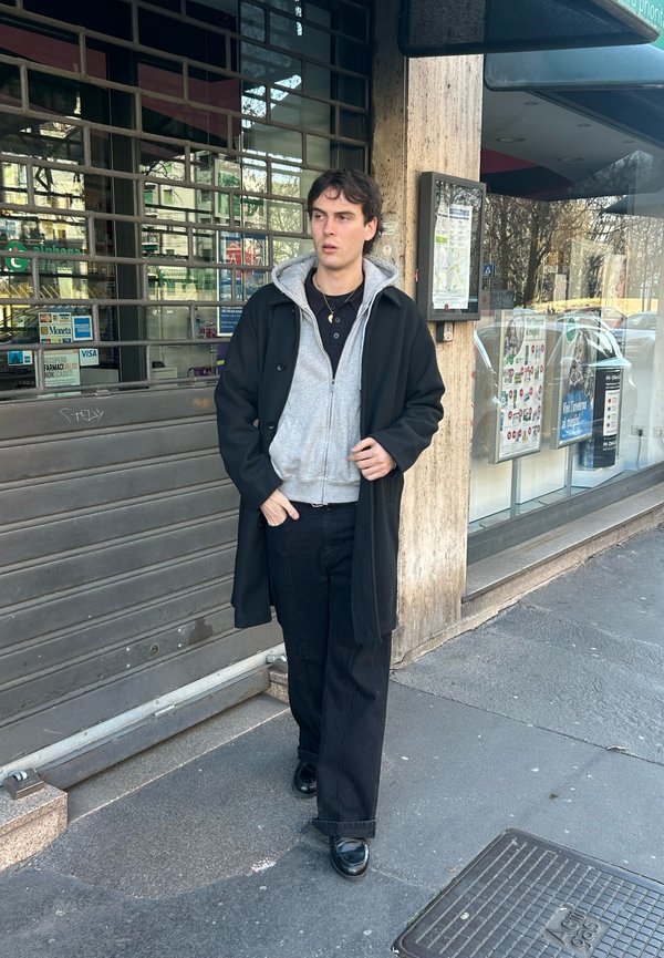 Tânăr în palton negru și hanorac gri, stând pe trotuar lângă un magazin închis cu oblon metalic și geam de sticlă.