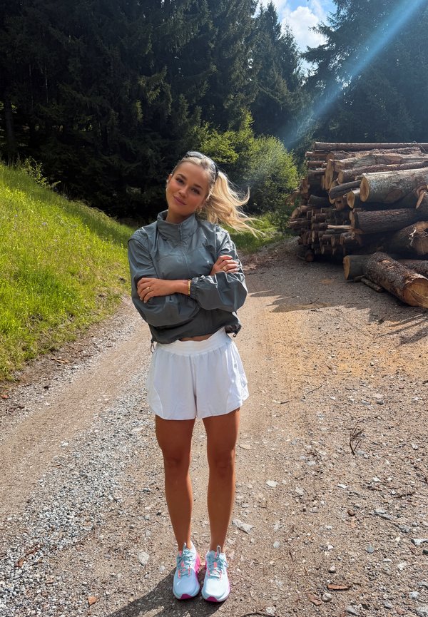 Joven mujer con ropa deportiva está de pie con los brazos cruzados en un sendero del bosque junto a troncos apilados bajo la luz del sol.