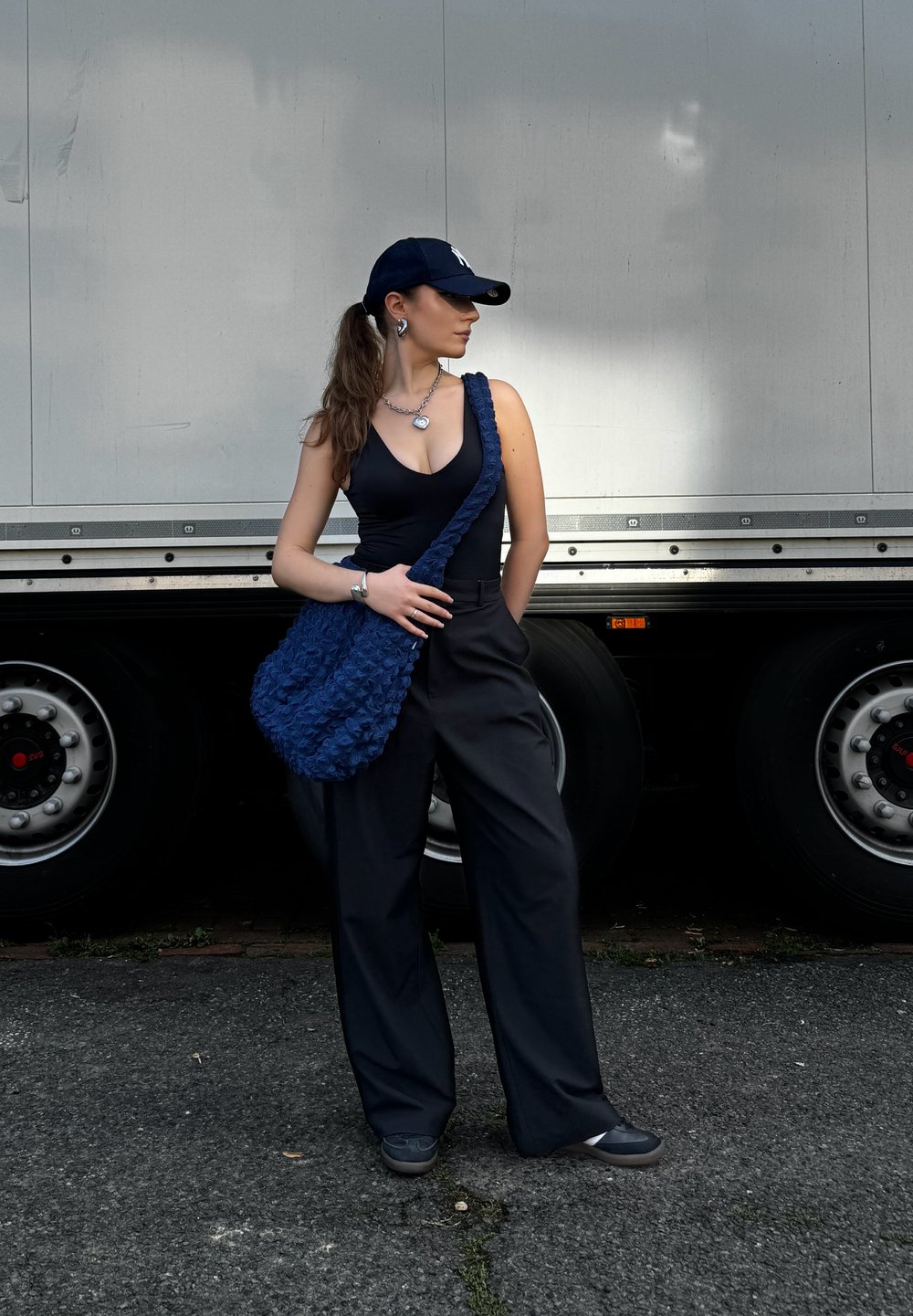Navy blue bag with textured surface, paired with a black bodysuit and wide-leg trousers. Accessories include a silver necklace and a cap.