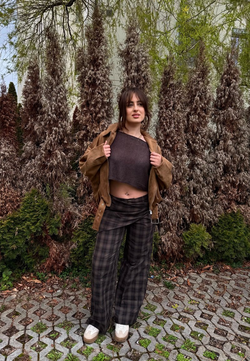 Jonge vrouw staat buiten op een geplaveide grond met gemengde groene en bruine dennenbomen achter zich, draagt een bruin jasje, een cropped top, geruite broek en witte schoenen.