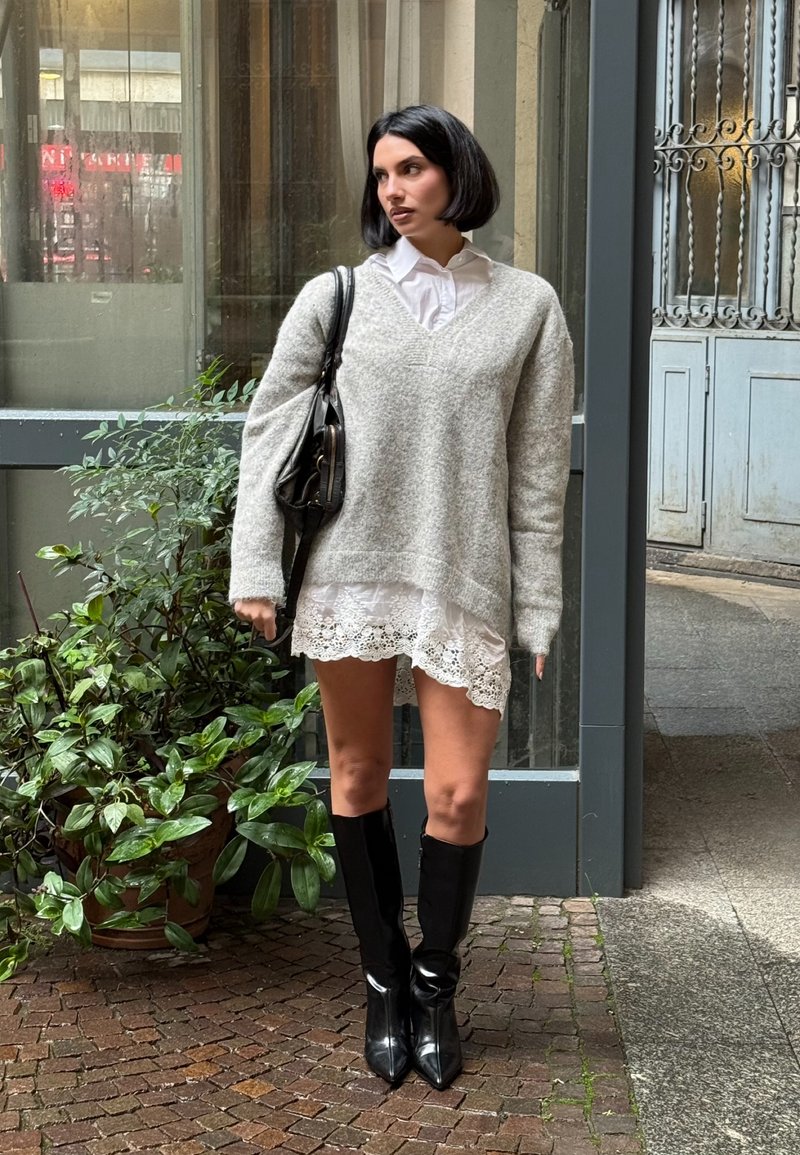 Camisola cinzenta oversized em malha, camisa branca com colarinho, vestido com renda nas bordas, botas pretas pontiagudas até ao joelho e mala preta. Plantas verdes ao fundo.
