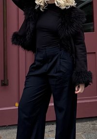 Čierna bunda z umelej kožušiny s textúrovanými manžetami, doplnená o čierne nohavice s vysokým pásom. Oblečenie má minimalistický dizajn s hladkým povrchom.