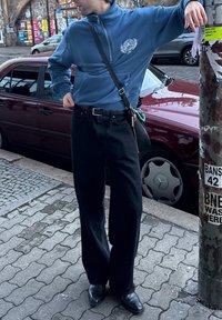 Μπλε φούτερ με φερμουάρ και λογότυπο, μαύρα παντελόνια σε ευθεία γραμμή και μαύρα παπούτσια με μυτερή μύτη, στέκεται κοντά σε μια κόκκινη αυτοκίνητο και σε μια κολώνα καλυμμένη με γκράφιτι.