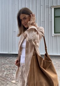 Egy barna vállig érő hajú nő haját igazítja, csíkos bézs ruhában, fehér felsőben, hajgumi van a hajában, és egy nagy barna bőr táskával sétál a szabadban.