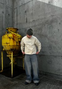 Light beige sweatshirt with a graphic print, loose blue jeans, and a green beanie. An aged yellow industrial container is in the background.