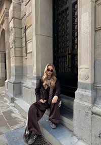 Brūnas plašas bikses ar baltām svītrām, melns tops un saulesbrilles. Fonā redzama akmens arhitektūra un melnas dzelzs vārti.