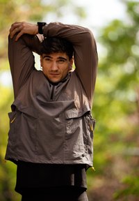 Tumean juustega mees, kes kannab pruunist jakki, venitab käsi pea kohal väljas, rohelise lehestiku uduses taustas.