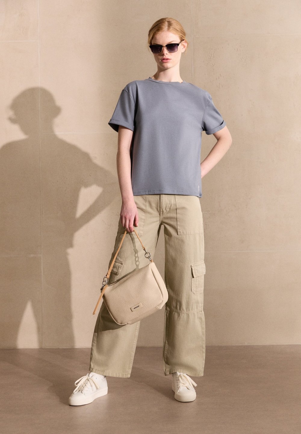 T-shirt in cotone grigio con maniche corte, pantaloni cargo color khaki, sneaker bianche e una borsa a tracolla beige con dettagli decorativi.