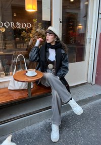 Veste en cuir noire, haut blanc, pantalon large gris et baskets blanches épaisses. Écharpe et béret soulignent la tenue. Café sur une table en bois.
