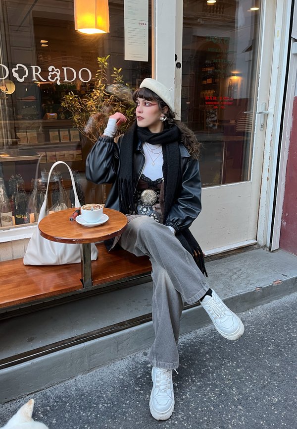 Schwarze Lederjacke, weißes Oberteil, graue weite Hose und weiße klobige Sneakers. Schal und Barett verleihen dem Outfit Akzente. Kaffee auf einem Holztisch.